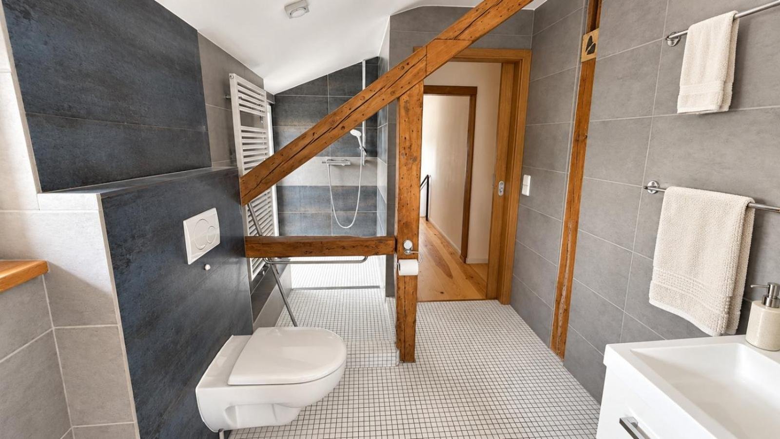 Bad mit Dusche, Wand-WC, grauen Fliesen und sichtbarem Holzbalken